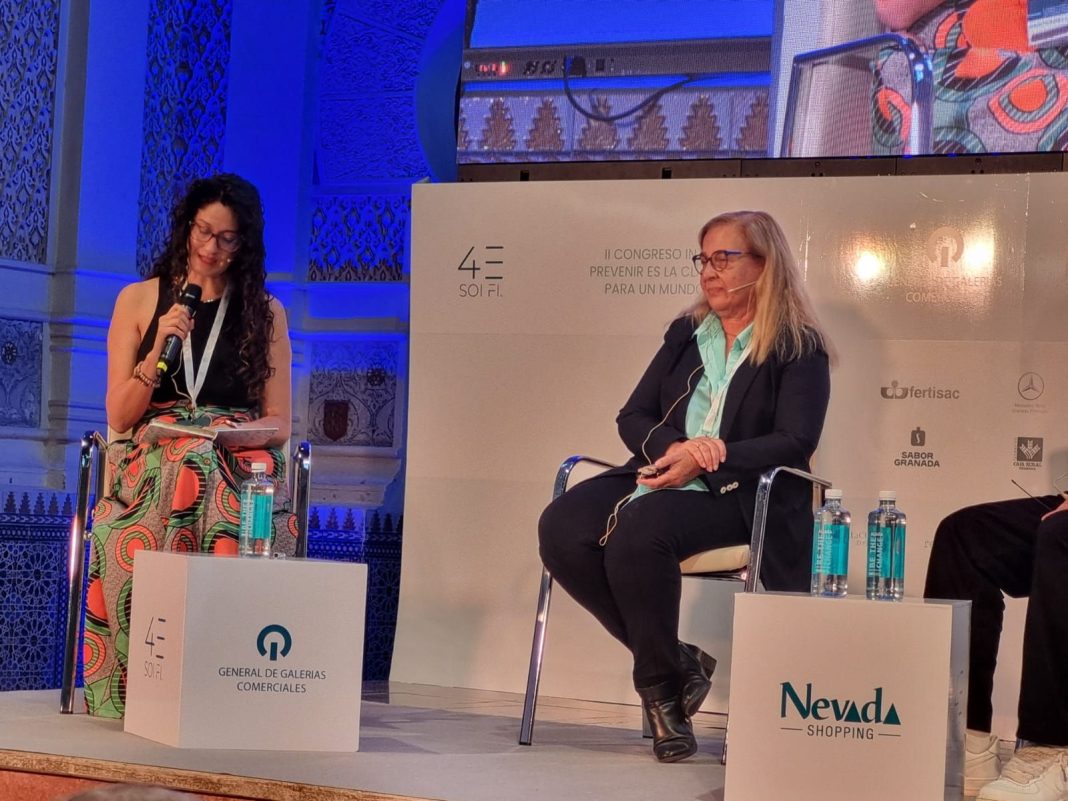 Dos mujeres sentadas participan en una charla durante un evento corporativo en un escenario iluminado con tonos azules. La moderadora, a la izquierda, lleva gafas, viste un top negro y una falda larga con un diseño colorido de patrones geométricos. Ella sostiene un micrófono y lee un documento en sus manos. A su lado derecho, una participante con cabello rubio largo y liso lleva una chaqueta oscura sobre una camisa verde clara, mientras escucha atentamente con un micrófono de diadema. En el fondo se observan decoraciones con mosaicos en relieve. En el escenario, destacan dos cubos con logotipos: uno de 
