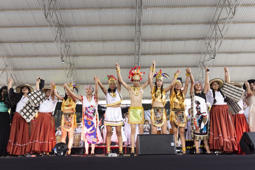 La imagen muestra a un grupo de personas sobre un escenario techado, todas tomadas de la mano con los brazos levantados en señal de celebración o finalización de una presentación. Están vestidas con trajes coloridos y tradicionales que parecen representar diversas culturas indígenas latinoamericanas. Algunas llevan faldas largas y sombreros de ala ancha, mientras otras están vestidas con trajes dorados, plumas de colores en la cabeza y joyería que evoca símbolos prehispánicos. El ambiente parece festivo, con un sentido de unidad y diversidad cultural.