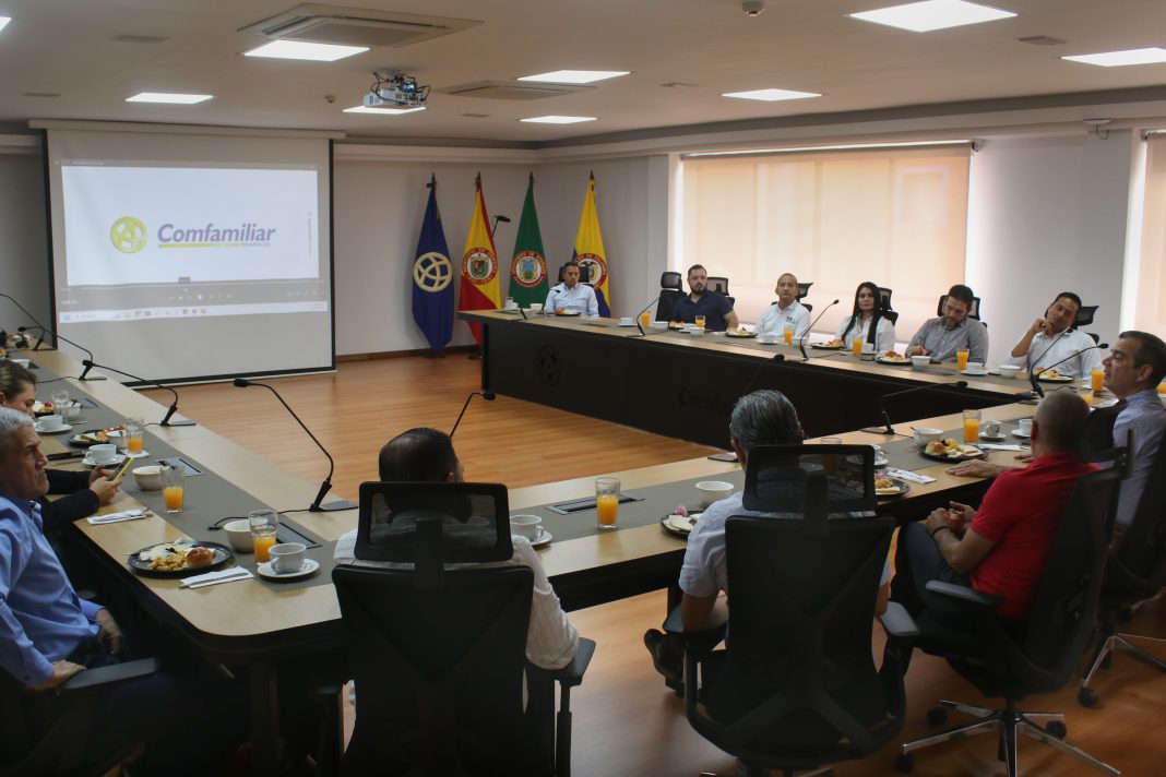 La imagen muestra una sala de reuniones con una mesa grande en forma de U, donde varias personas están sentadas participando en una reunión. En el centro de la sala hay una proyección en la pared que muestra el logotipo de 