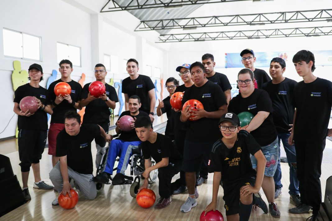 La imagen muestra a un grupo de personas jóvenes, la mayoría con camisetas negras, posando para una foto en una bolera. Algunos de los participantes están de pie, mientras que otros están agachados o en sillas de ruedas. Todos sostienen bolas de boliche de diferentes colores. Hay un ambiente alegre y de compañerismo entre los presentes. Se observa que algunos de los jóvenes tienen discapacidad, pero están participando activamente en la actividad deportiva, lo que refleja inclusión y diversión.