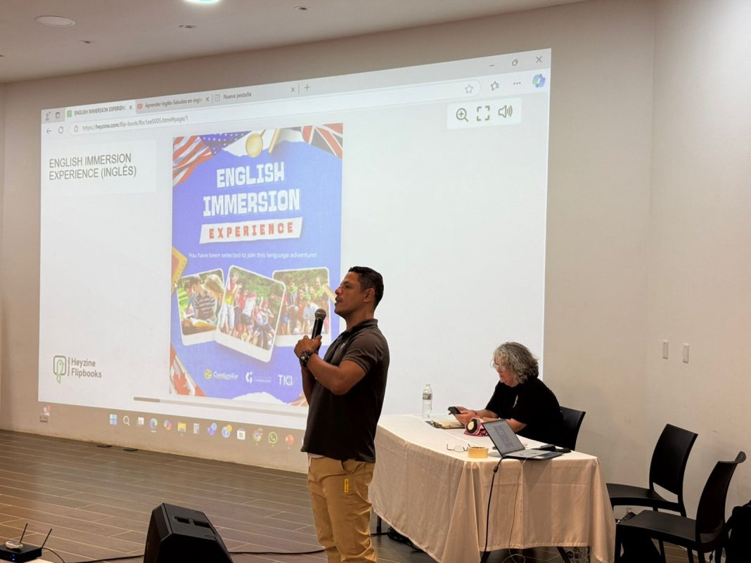 La imagen muestra a una persona de pie, hablando con un micrófono frente a una pantalla de proyección en una sala de conferencias. La pantalla proyecta un contenido titulado 