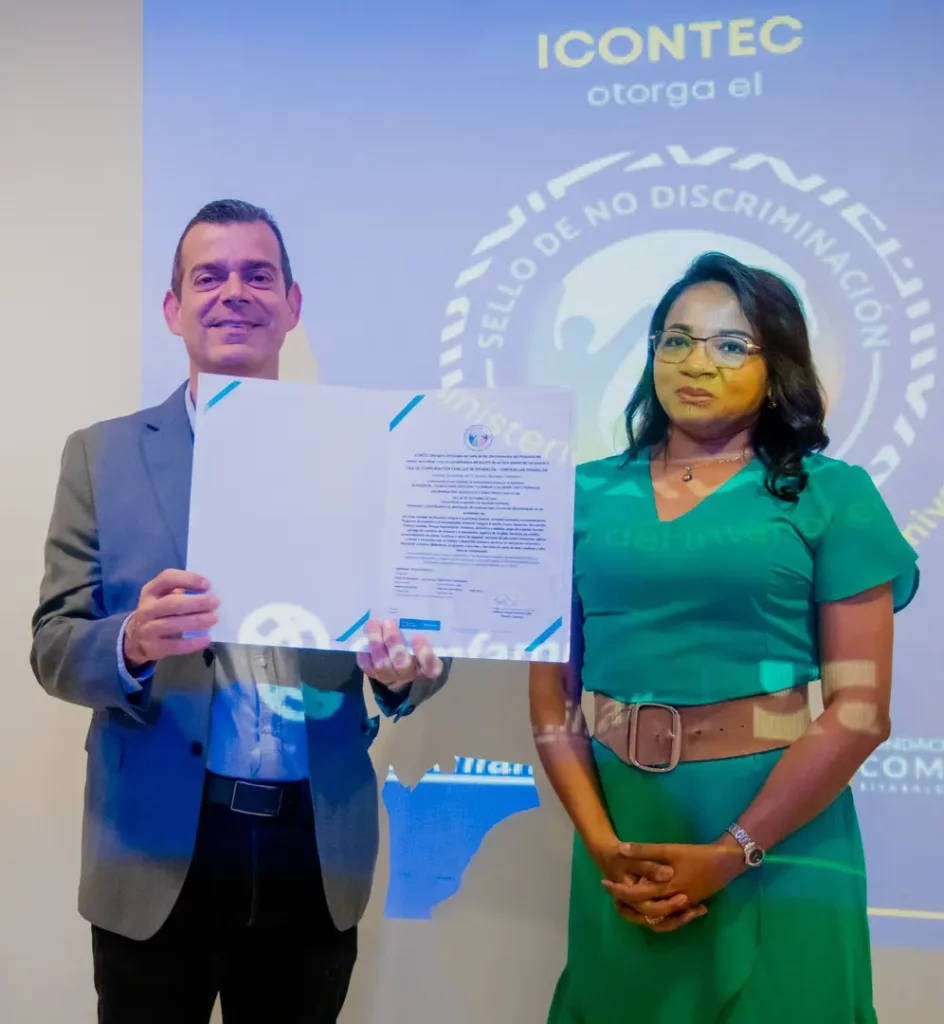 Un hombre y una mujer posan durante la entrega del Sello de No Discriminación otorgado por ICONTEC. El hombre, vestido con traje gris, sostiene el certificado abierto mientras sonríe. A su lado, la mujer, con un vestido verde, permanece de pie con expresión serena. Al fondo, una proyección en la pared muestra el logotipo del reconocimiento y el texto 'Sello de No Discriminación'.